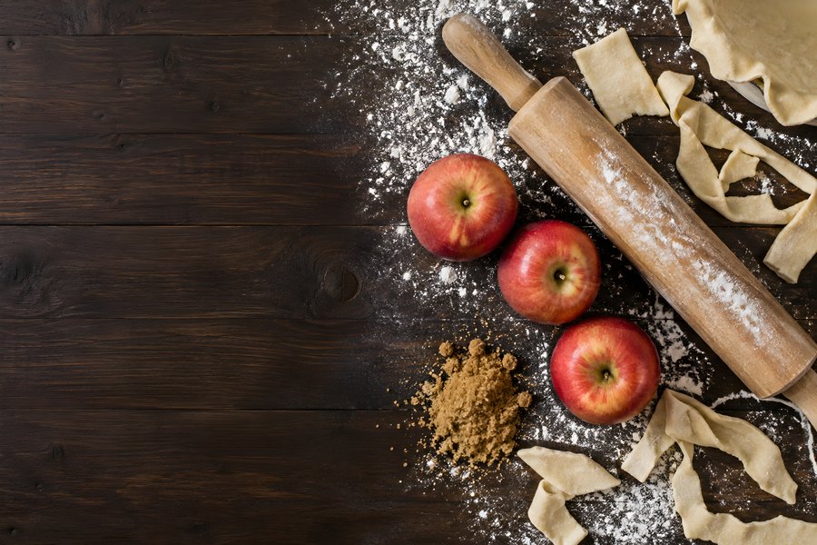 Rustic apple pie preparation with fresh apples, floured rolling pin and pastry dough on a dark wood table