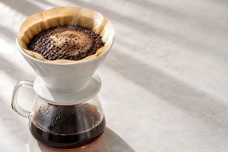 White ceramic pour-over dripper with blooming coffee grounds and rising steam on a glass carafe, sunlit concrete surface
