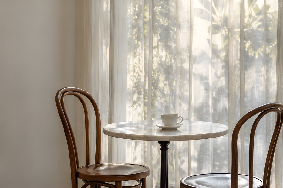 Empty cafe corner with a marble bistro table, two bentwood chairs and sheer curtains lit by warm window light