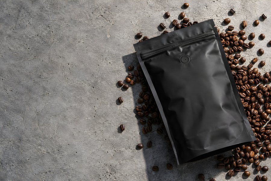 Blank matte black coffee pouch with degassing valve and scattered roasted beans on grey concrete, flat lay with copy space