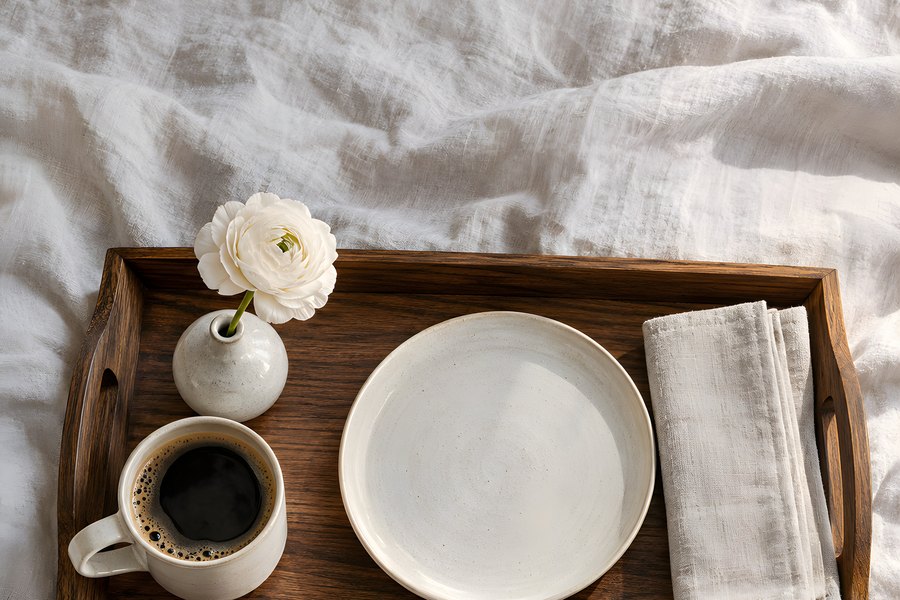 Wooden breakfast tray with black coffee, empty stoneware plate, folded linen napkin and a white ranunculus on a rumpled bed