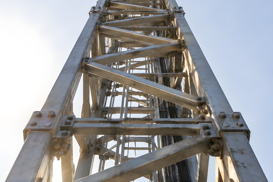 Low-angle view looking up at a tower crane's steel lattice mast and jib against a clear blue sky with sun flare