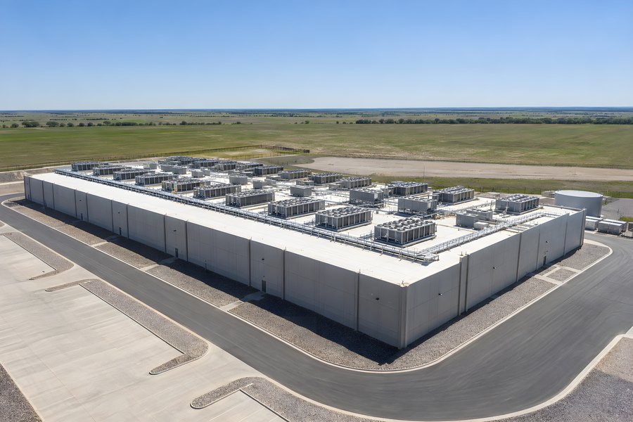 Aerial view of a windowless data center building with rooftop cooling units beside a water tank on open grassland