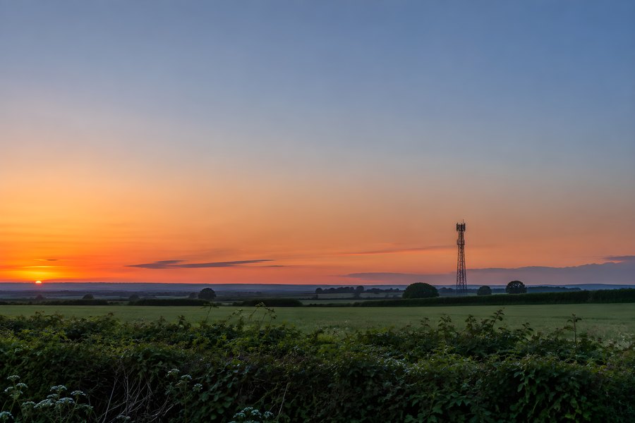Lattice telecom tower silhouetted against a vivid sunset sky over green farmland with a wildflower hedgerow in the foreground