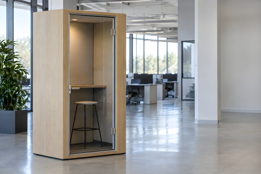 Empty wood-veneer acoustic phone booth with glass door and stool in a modern open-plan office