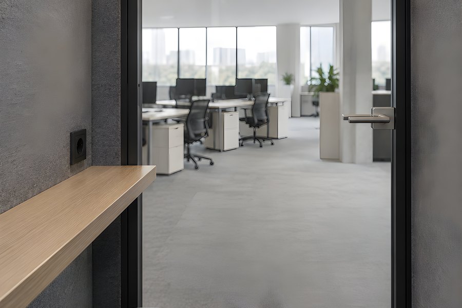 Empty acoustic phone booth with a wooden fold-down desk and stool opening onto a modern open-plan office with a city view