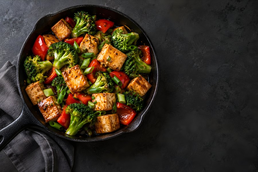 Vegan tofu and vegetable stir fry with broccoli and red pepper in a cast iron skillet, overhead view with copy space
