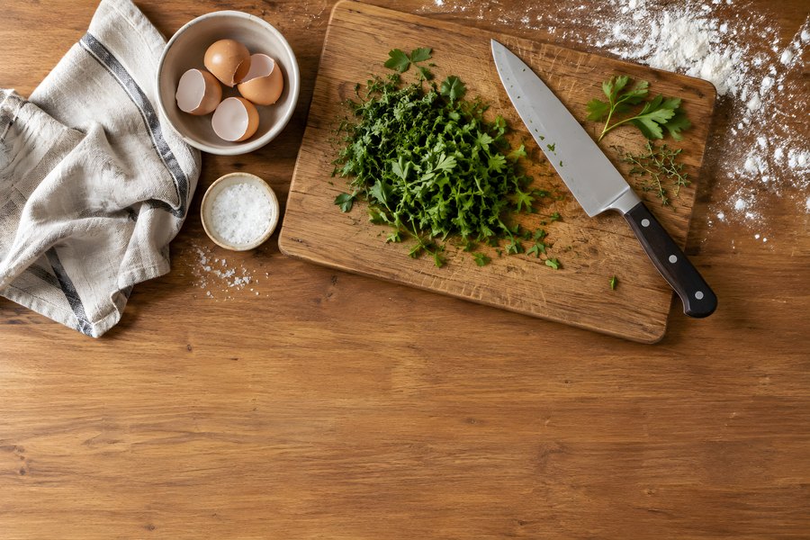 Wooden chopping board with fresh chopped parsley, chef's knife and eggshells on a rustic kitchen counter with flour dusting