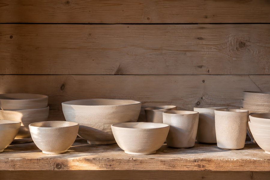Handmade unglazed ceramic bowls and cups arranged on a rustic wooden shelf in warm sunlight