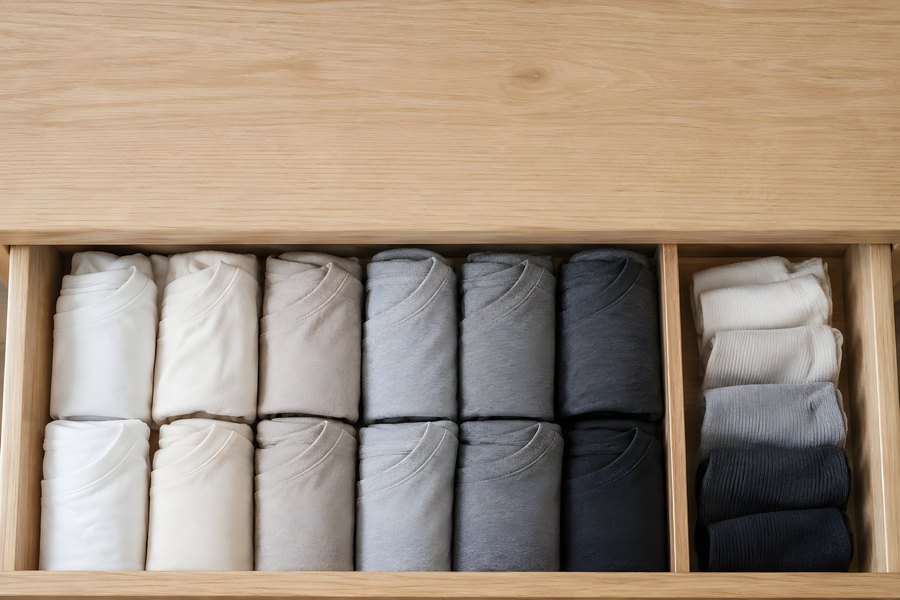 Folded t-shirts filed upright by tone in a wooden drawer beside a divided compartment of rolled clothing