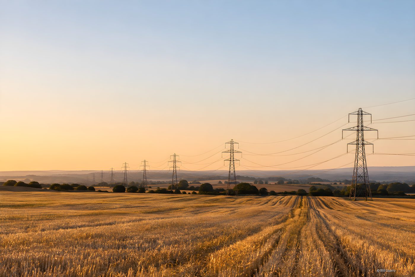 Row of steel lattice transmission pylons carrying overhead power lines across a harvested golden stubble field at dusk