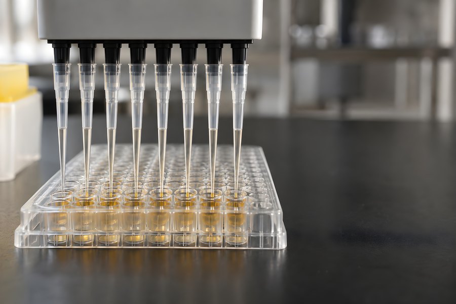 Multichannel pipette dispensing amber liquid into a 96-well microplate in a laboratory, close-up macro shot with no people