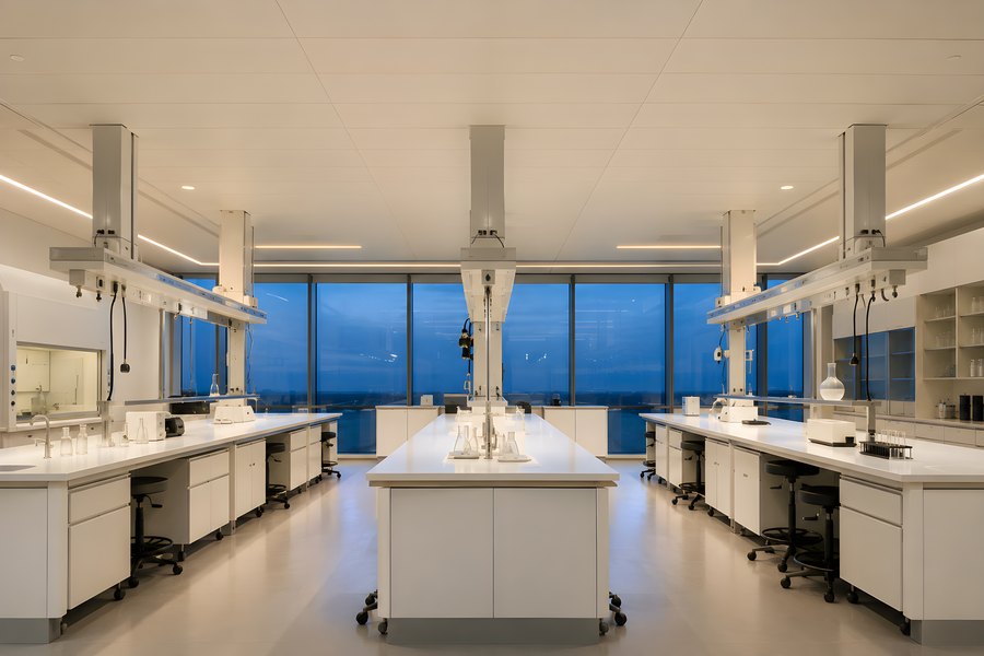 Empty modern laboratory with central island bench and long white worktables at dusk, floor-to-ceiling windows overlooking city lights