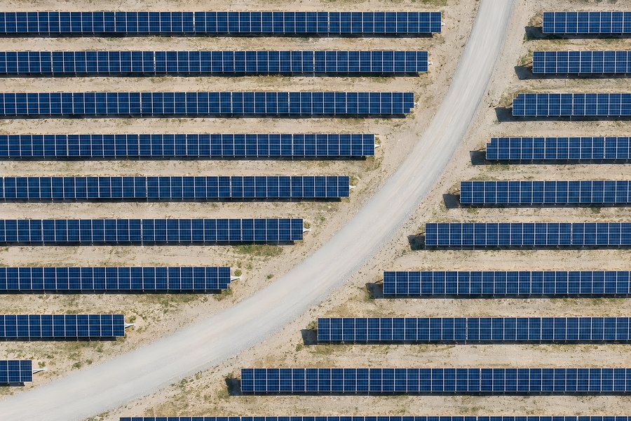 Aerial top-down view of utility scale solar farm with rows of blue photovoltaic panels beside a curved gravel road on arid land