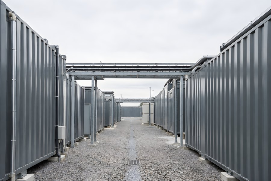 Battery energy storage system site with grey steel containers and an overhead pipe bridge along a gravel service aisle under overcast sky