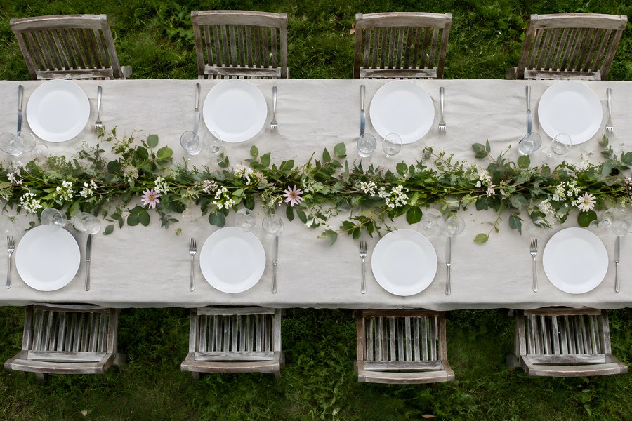 Overhead view of a long outdoor table set with white plates, linen cloth and a greenery garland runner, no people