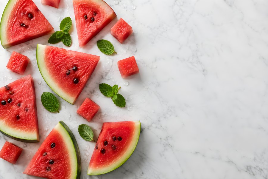 Overhead flat lay of watermelon slices, wedges and cubes with mint leaves and water droplets on marble with copy space