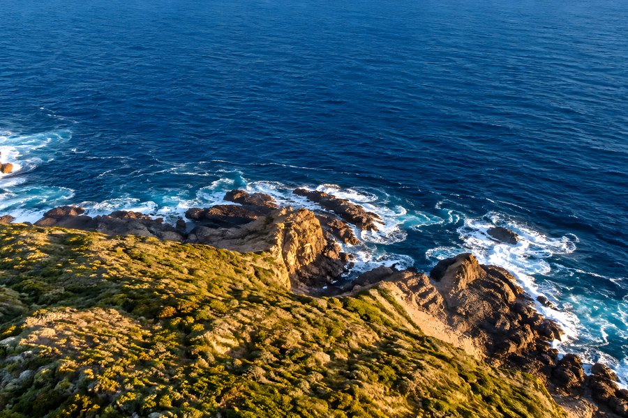 Aerial drone view of a rugged rocky coastline with ocean waves breaking against cliffs and a green headland at golden hour