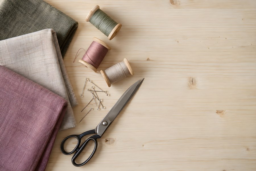 Overhead flat lay of sewing supplies with folded linen fabric, thread spools, pins and dressmaking shears on a wooden table