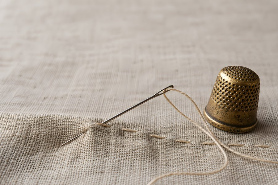 Threaded sewing needle stitching natural linen fabric next to a vintage brass thimble in a close-up craft still life