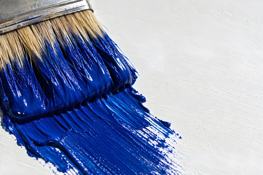 Close-up of a wide paintbrush loaded with bright blue paint dragging a glossy stroke across a white textured wall