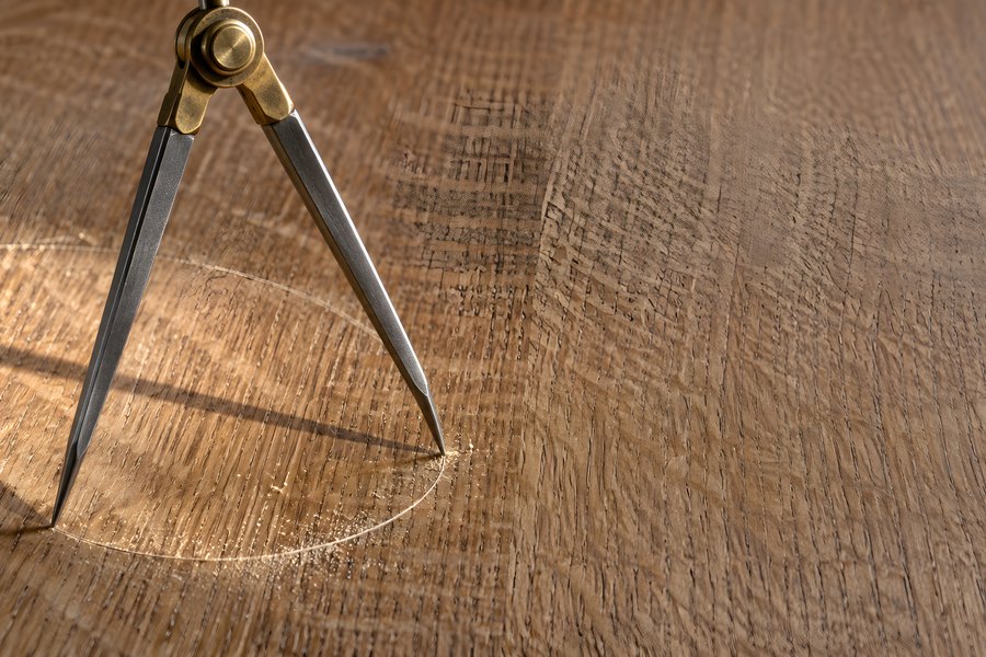 Brass and steel drafting dividers scribing a circle into oak wood grain, close-up marking and layout tool