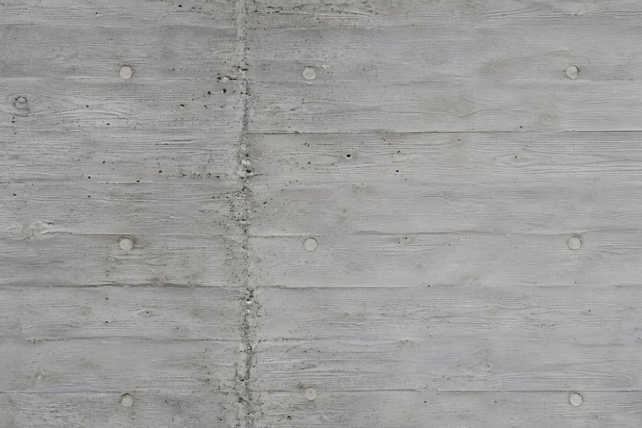 Board-formed raw concrete wall texture with round tie holes and a vertical panel joint, grey brutalist background