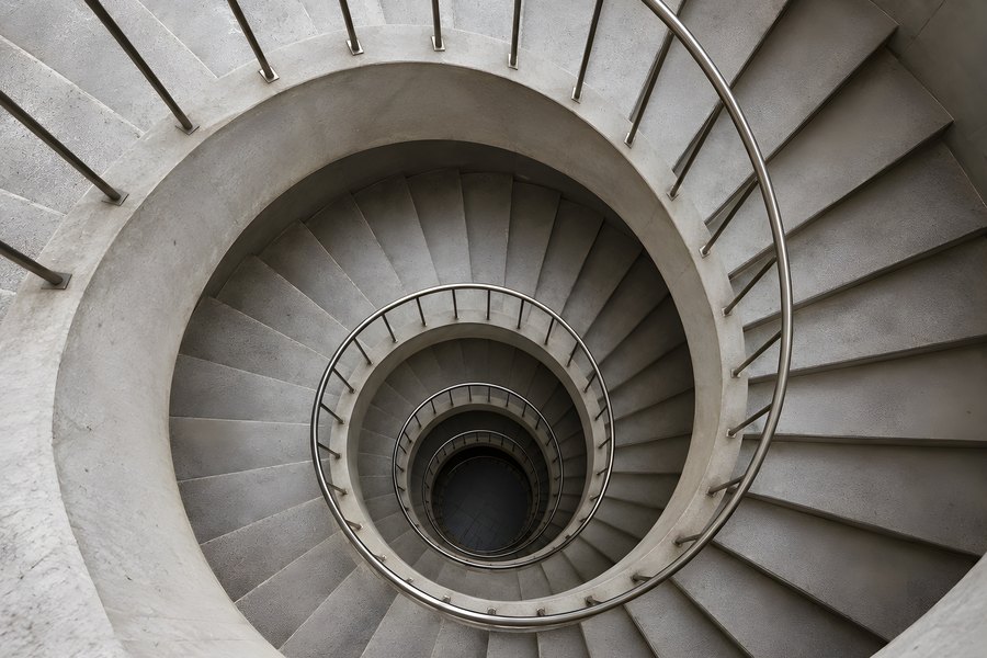 Spiral concrete staircase photographed from above with a curving stainless steel handrail winding down into a dark stairwell
