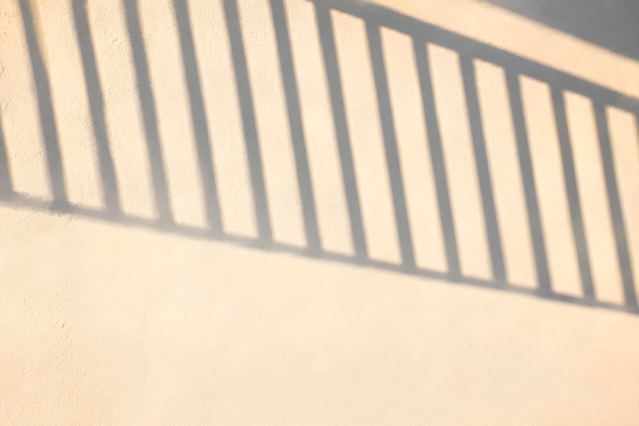 Diagonal railing shadow casting hard stripe patterns across a warm textured plaster wall